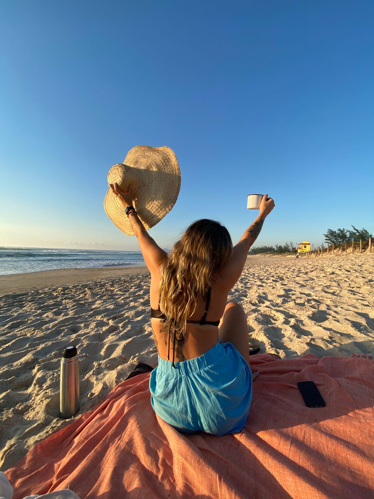 A woman on a beach blanket raising her sun hat toward a clear blue sky
