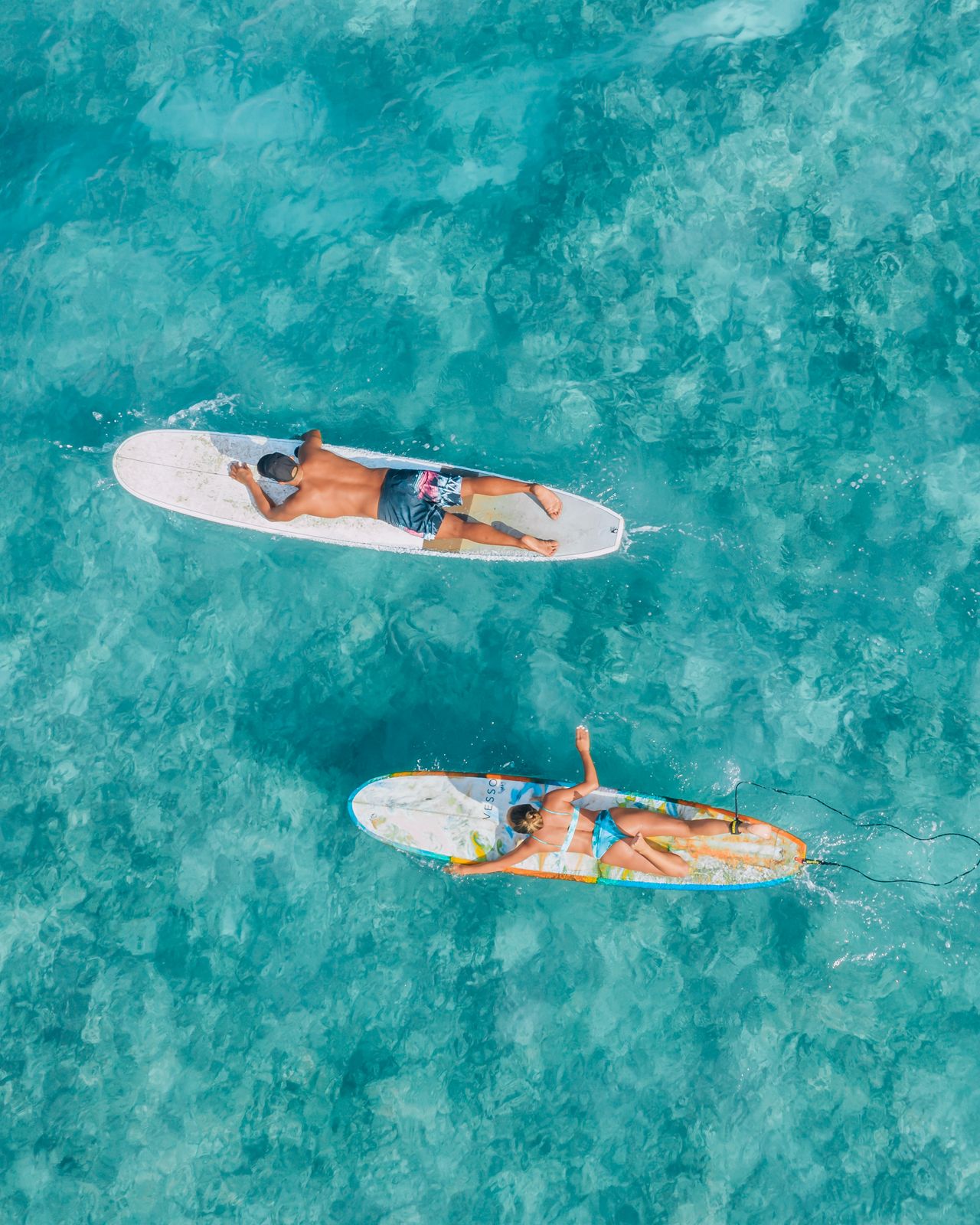 Two surfers paddling over clear turquoise water, seen from above