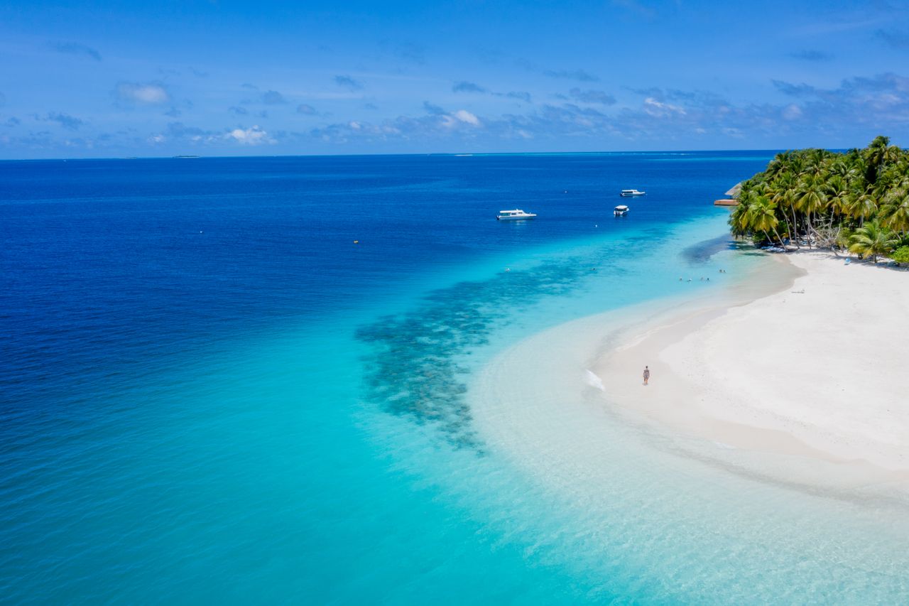 A white sand beach curving into turquoise water, with palms and boats offshore