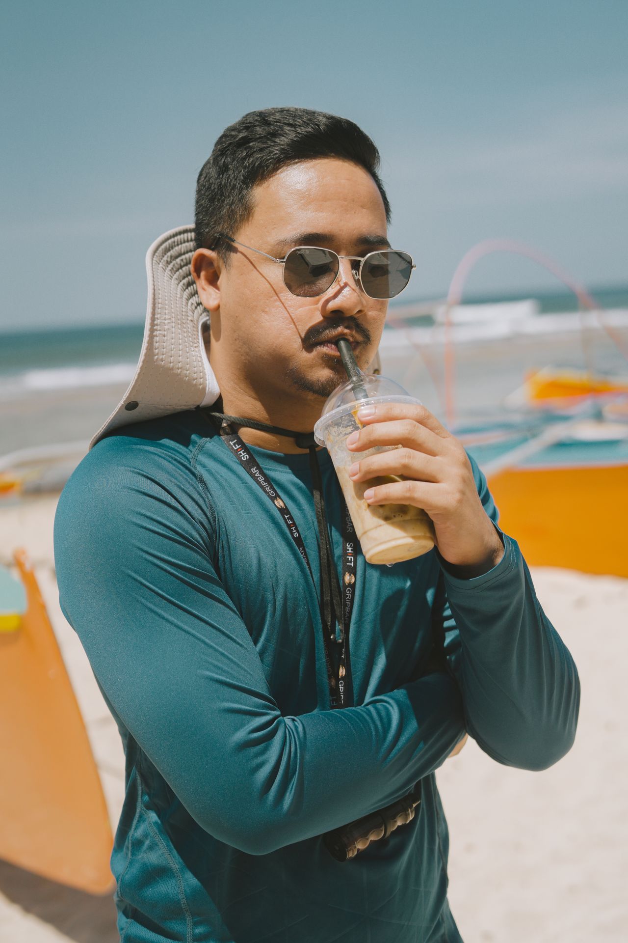 Ilyas Rasheed in sunglasses with an iced coffee on a beach lined with outrigger boats