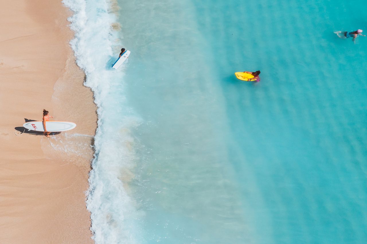 Aerial view of a surfer carrying a board into turquoise surf beside a sandy beach