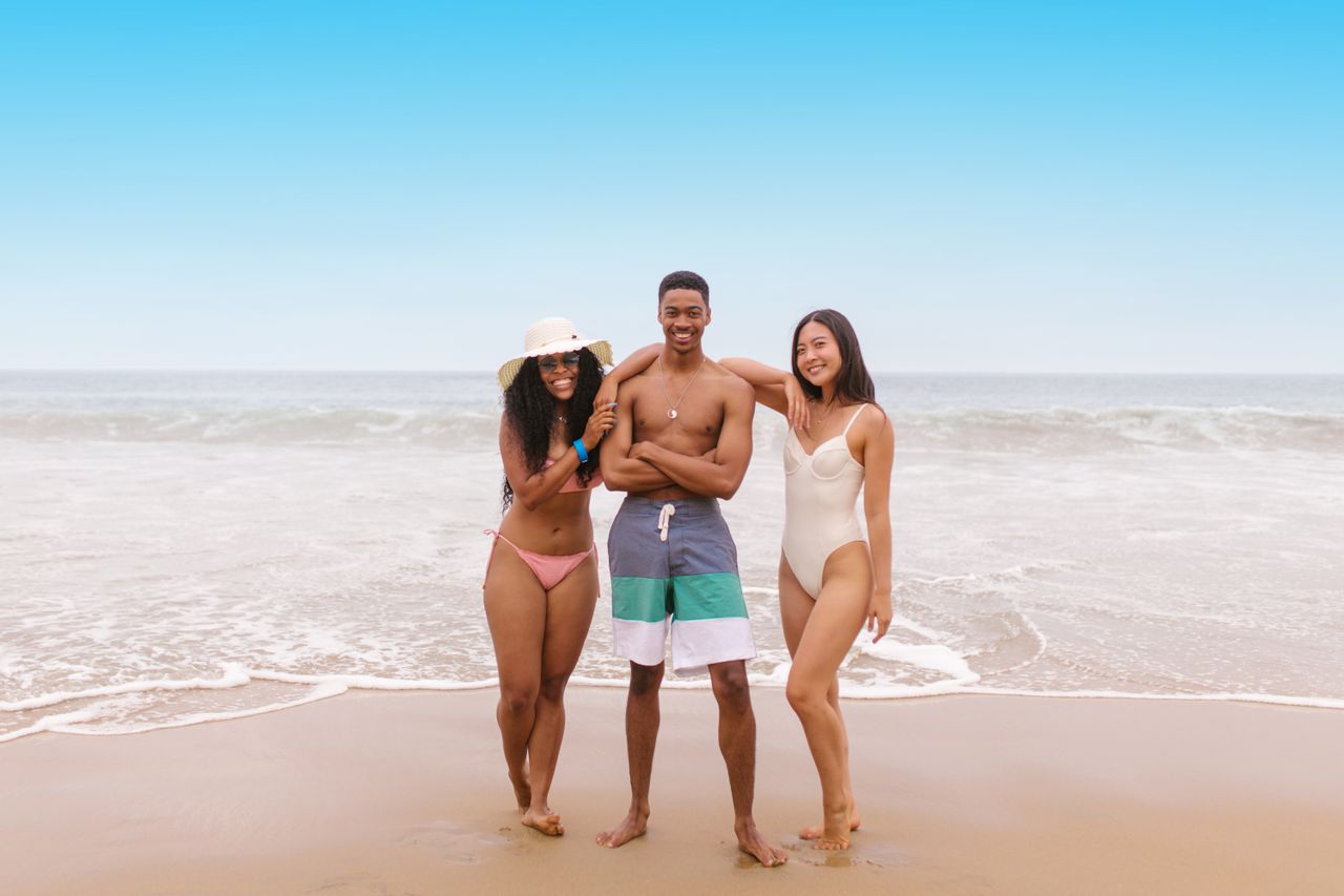 Three smiling hosts standing together at the water's edge on a sunny beach