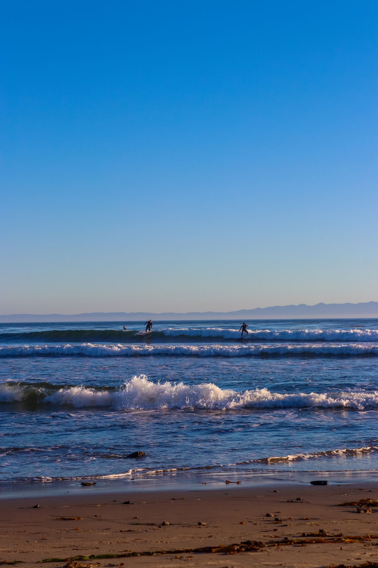 Clear blue sky over rolling surf, with surfers waiting for waves beyond the beach