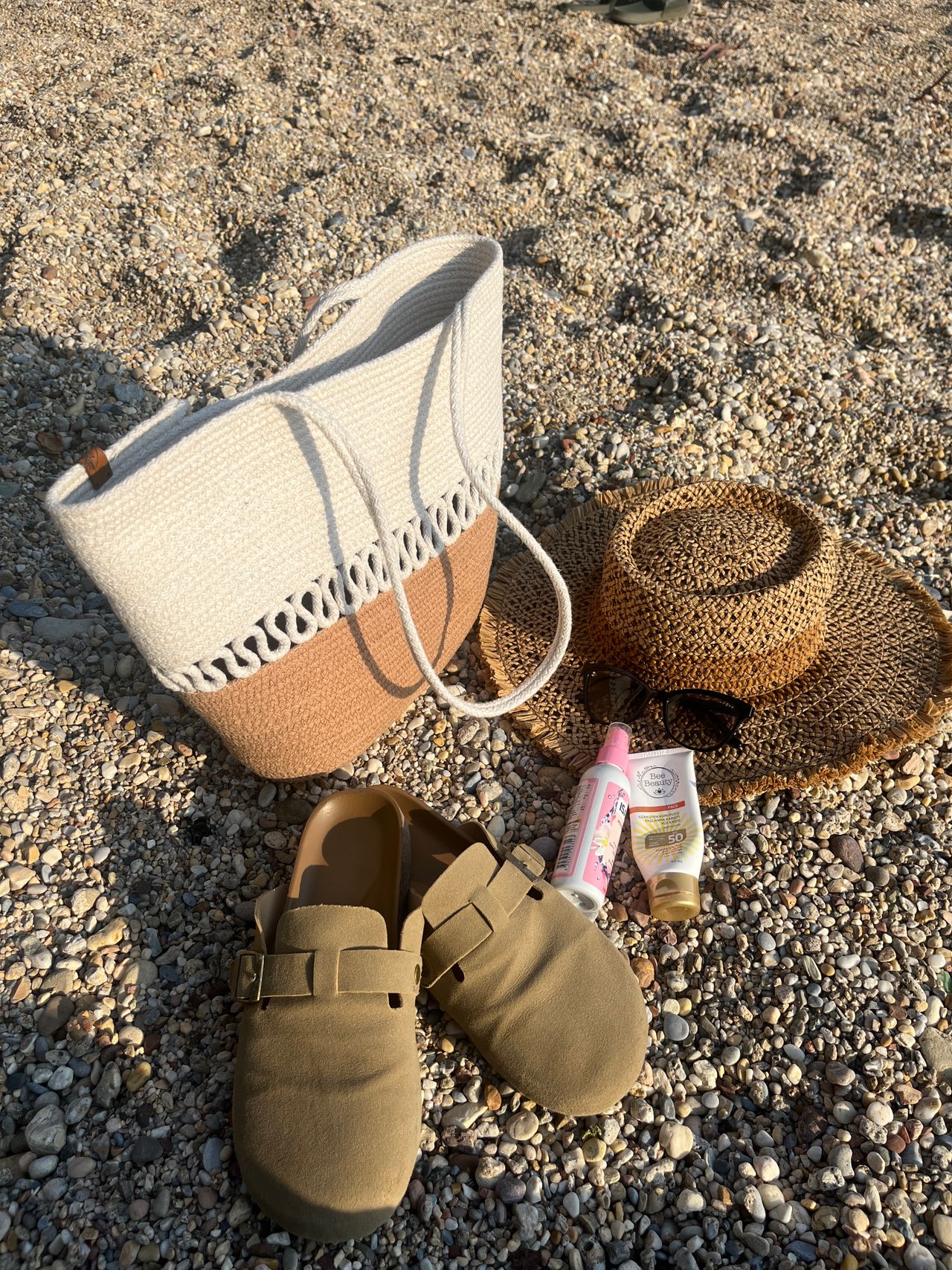 A woven beach bag, straw hat, sunglasses, sunscreen and suede sandals laid out on the sand