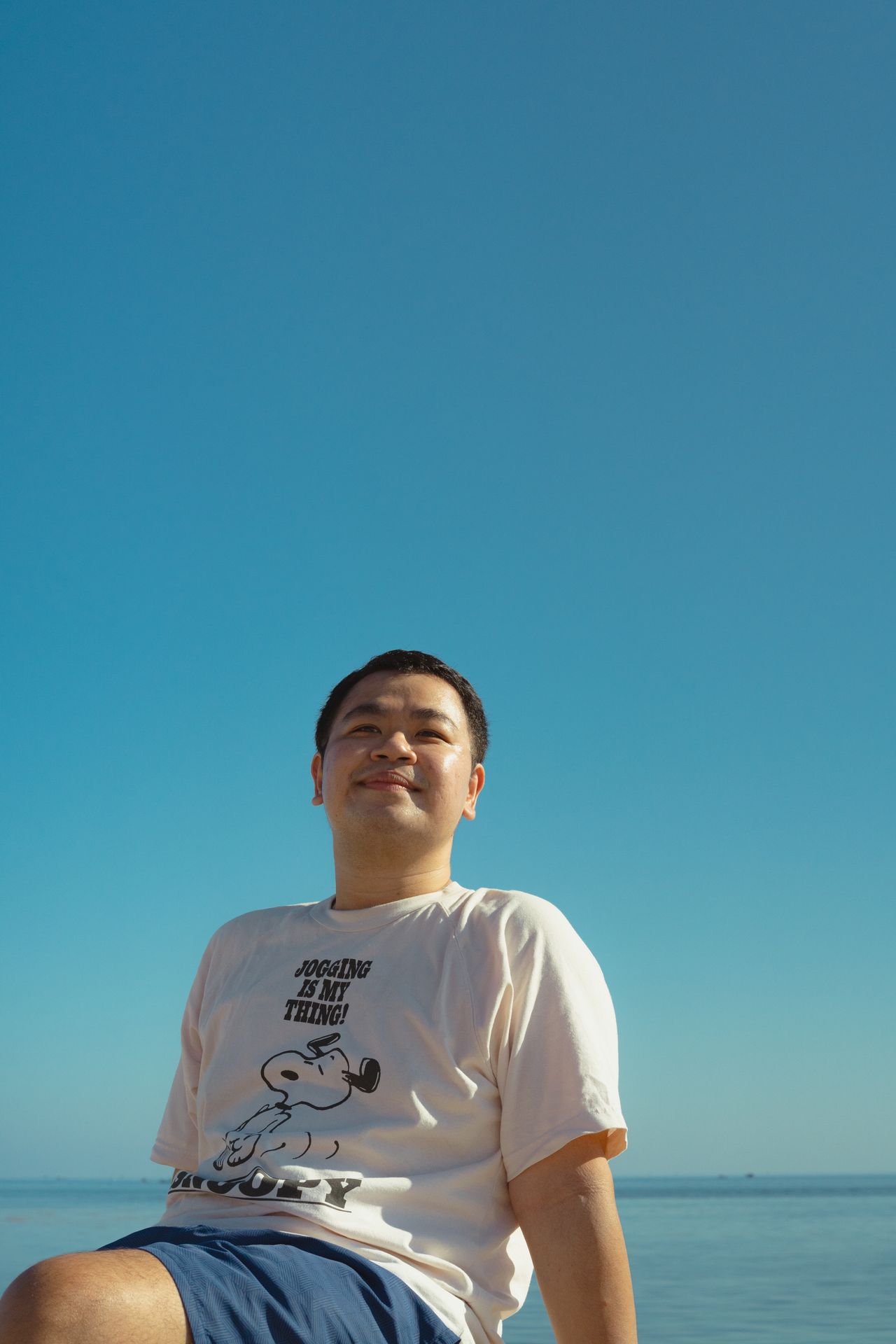 A smiling guest in a white T-shirt sitting by the sea under a clear blue sky